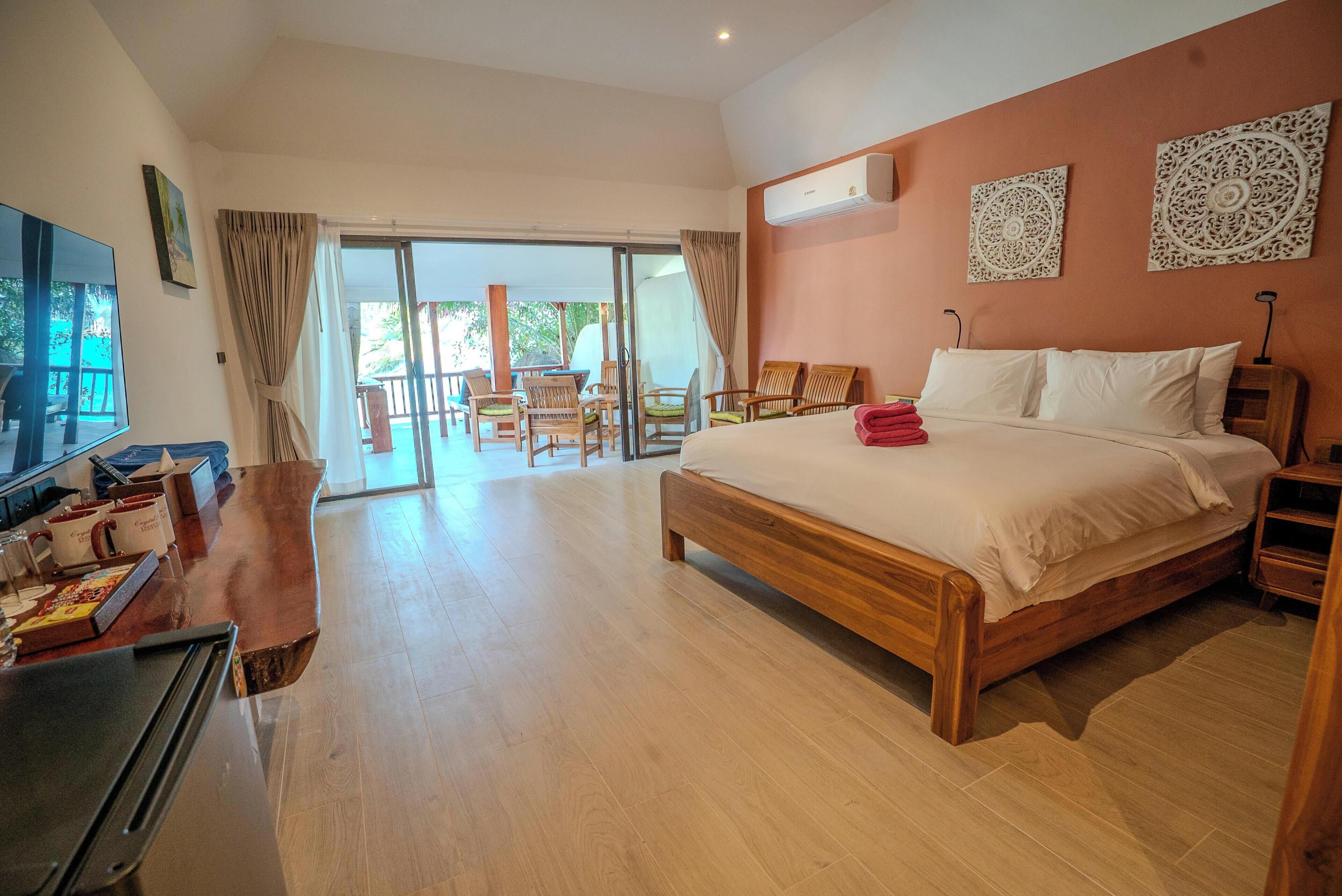 A spacious bedroom with a large bed, a wooden desk, and a TV. There is a balcony with chairs and a table.
