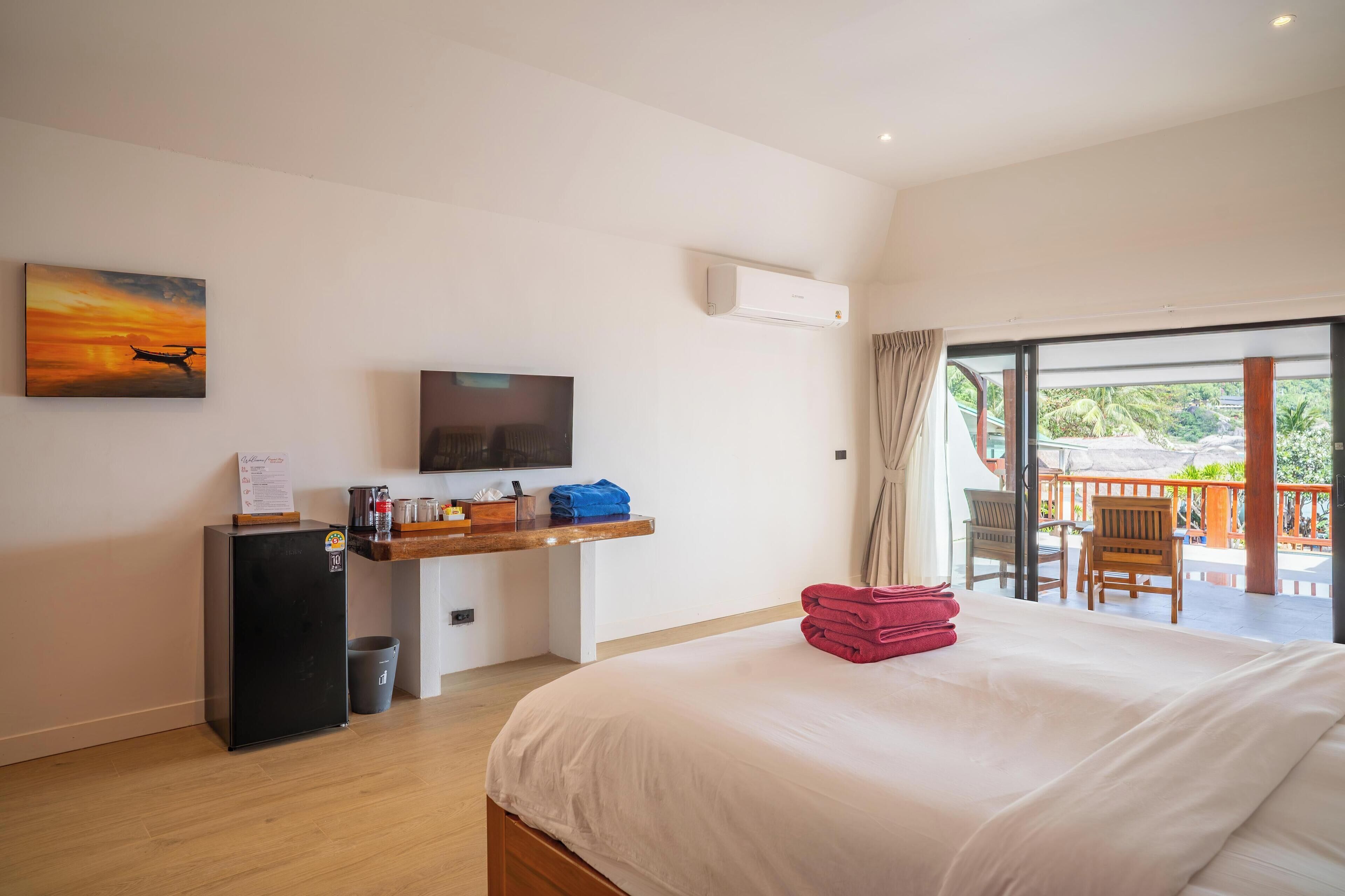 A hotel room with a bed, a desk with a flat-screen TV, a small refrigerator, and a balcony with outdoor seating.