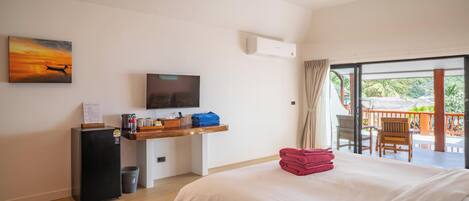 A hotel room with a bed, a desk with a flat-screen TV, a small refrigerator, and a balcony with outdoor seating.