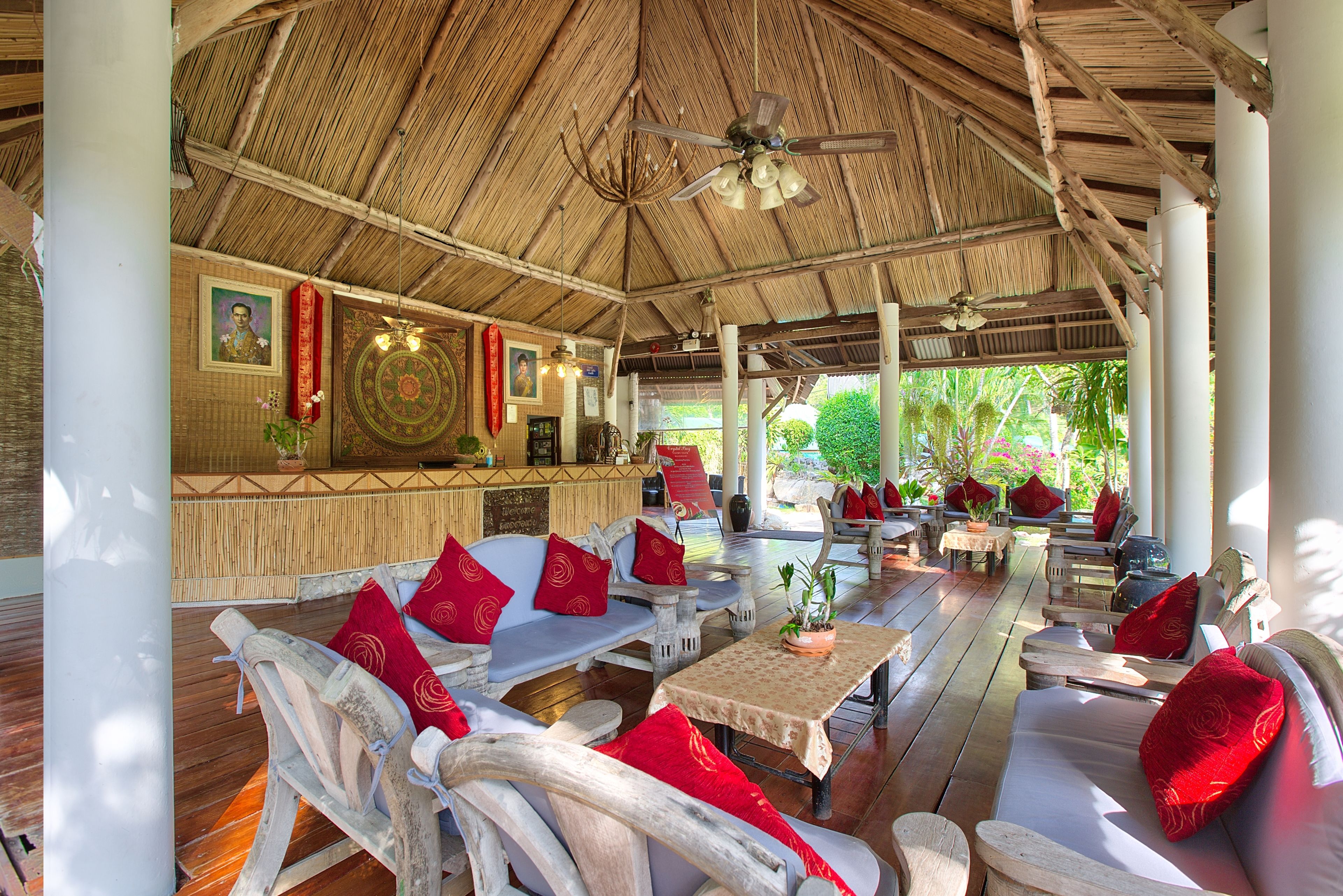 A covered outdoor seating area with wooden furniture, red cushions, and a central table with a potted plant.