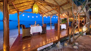 Un restaurante en la playa con terraza, techo de paja e iluminación con guirnaldas.