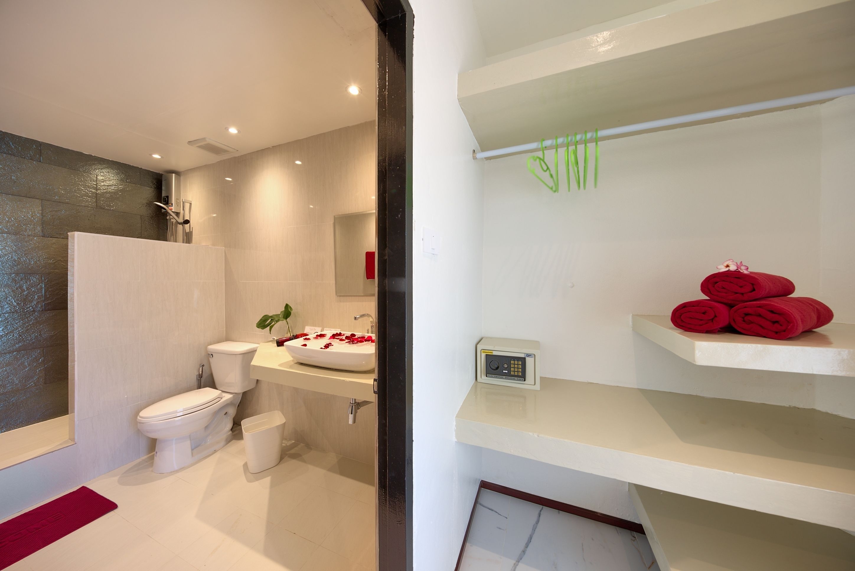 A modern bathroom with a toilet, sink, and shower. There are red towels on a shelf and a small plant on the sink.