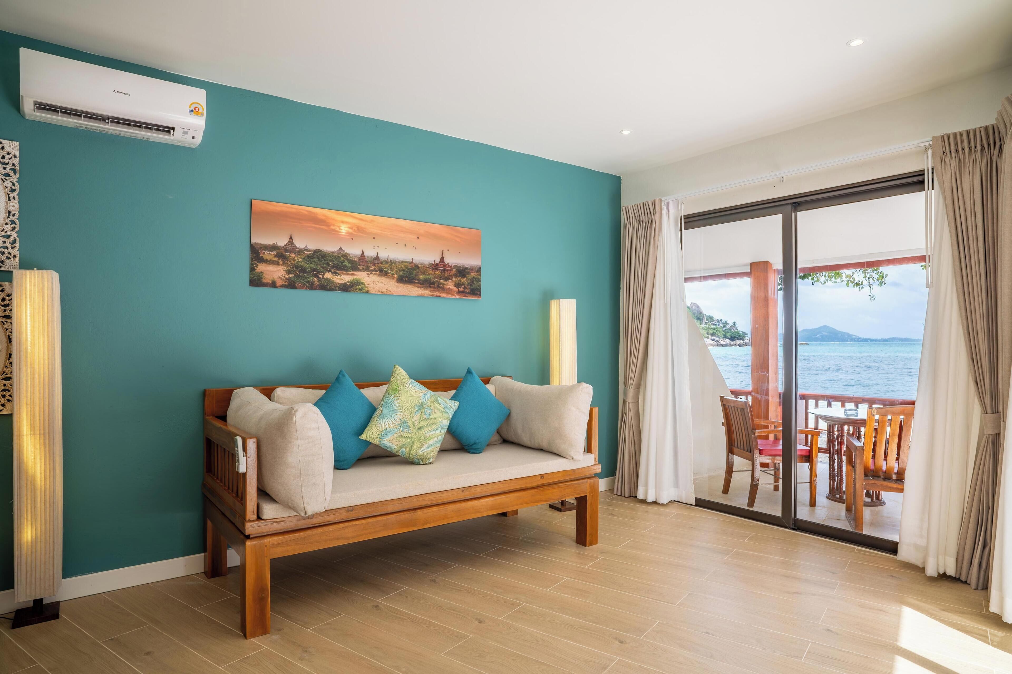 A room with a teal wall, a wooden sofa with cushions, a painting of a sunset, and a view of the sea through a glass door.