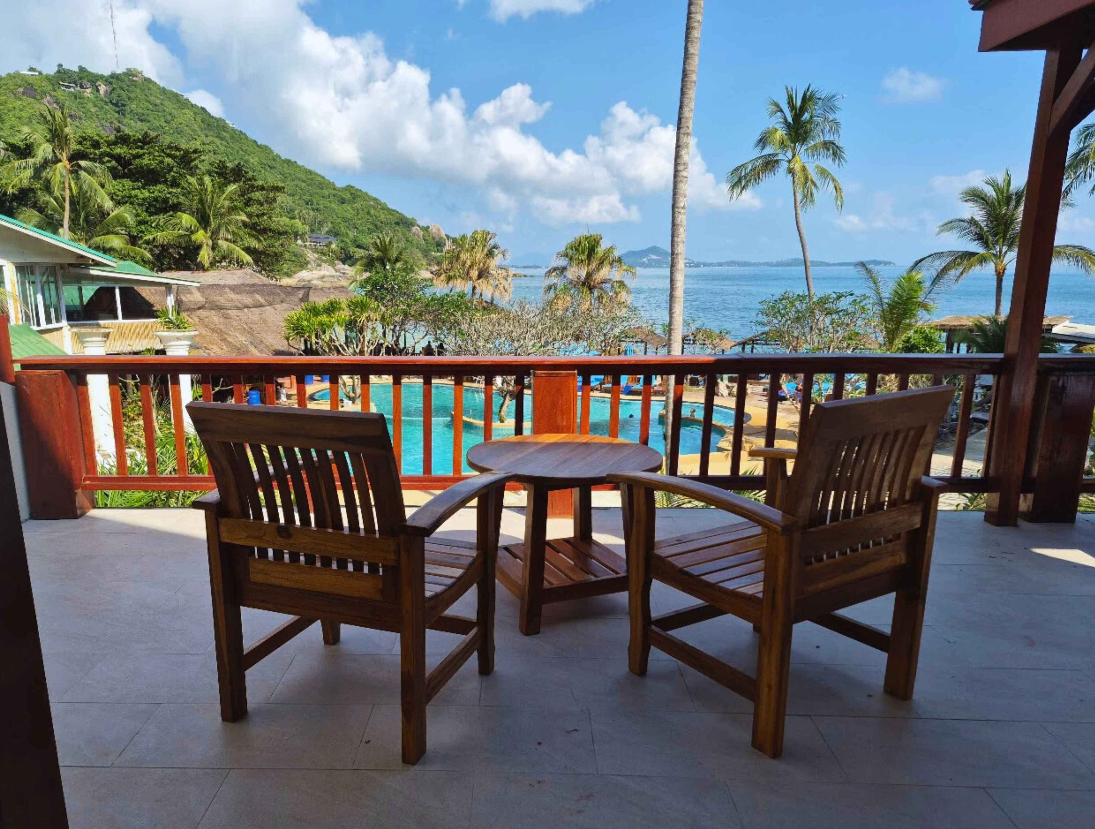 A wooden deck with two chairs and a table, overlooking a pool and a beach with palm trees and a mountain in the background.