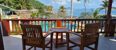 A wooden deck with two chairs and a table, overlooking a pool and a beach with palm trees and a mountain in the background.