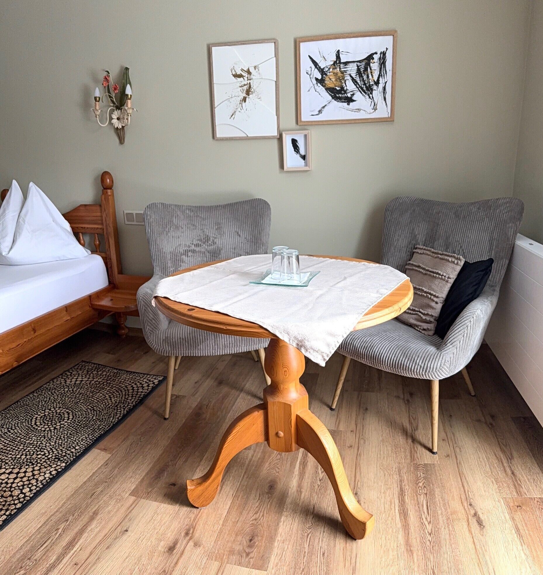 Une pièce comprenant un lit, deux chaises, une petite table recouverte d’une nappe blanche et deux œuvres d’art encadrées accrochées au mur.