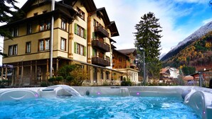 A hot tub with a mountainous backdrop, surrounded by traditional multi-story buildings with balconies.