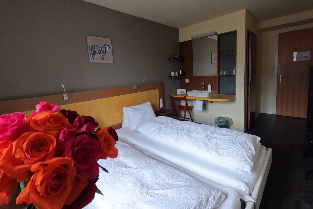 Une chambre d’hôtel avec un lit, un bureau, une chaise, un lavabo et un miroir. Un vase rempli de fleurs est posé sur le lit.