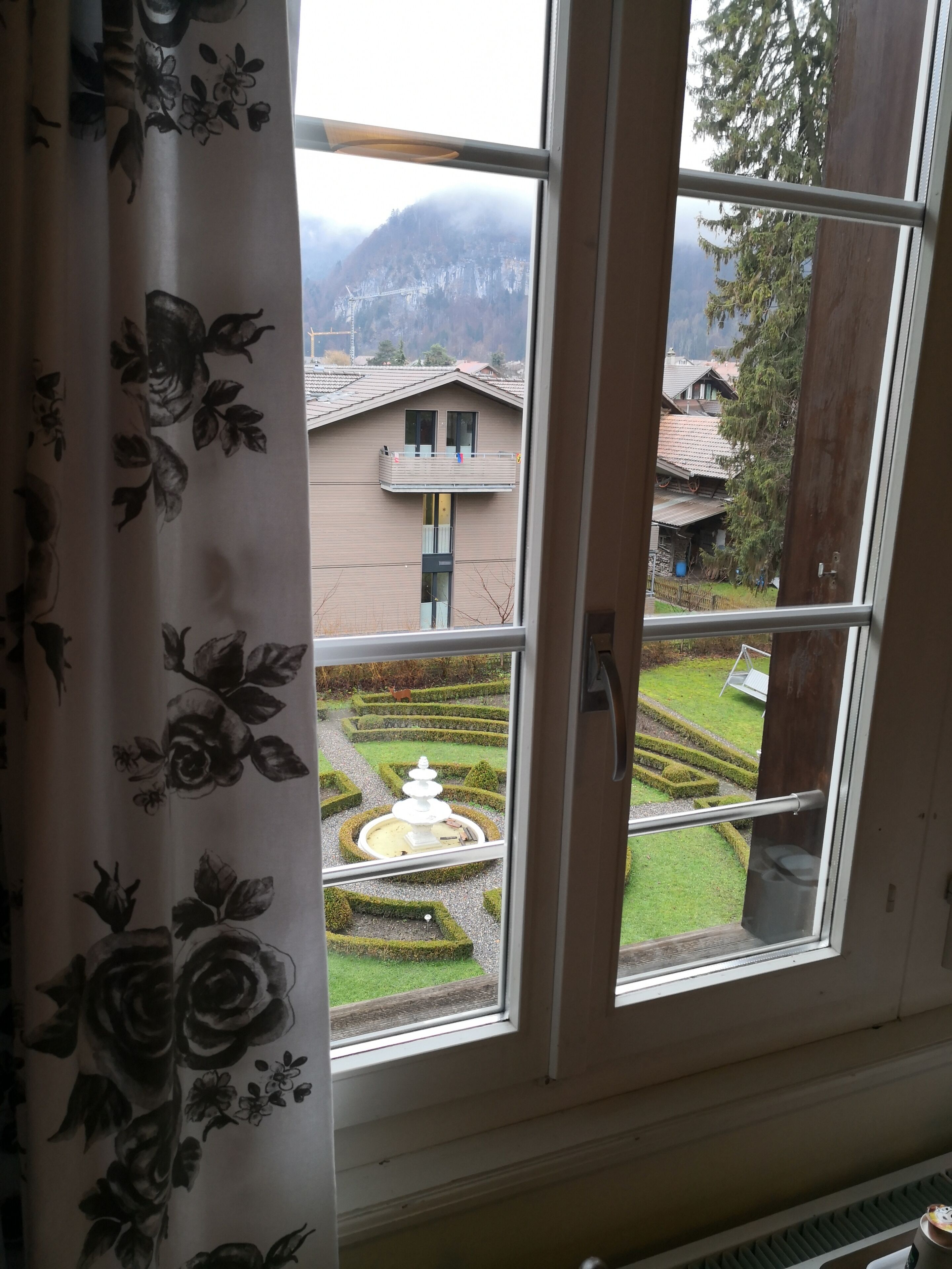 A room with a window overlooking a garden with a fountain, a balcony, and a view of a mountain.