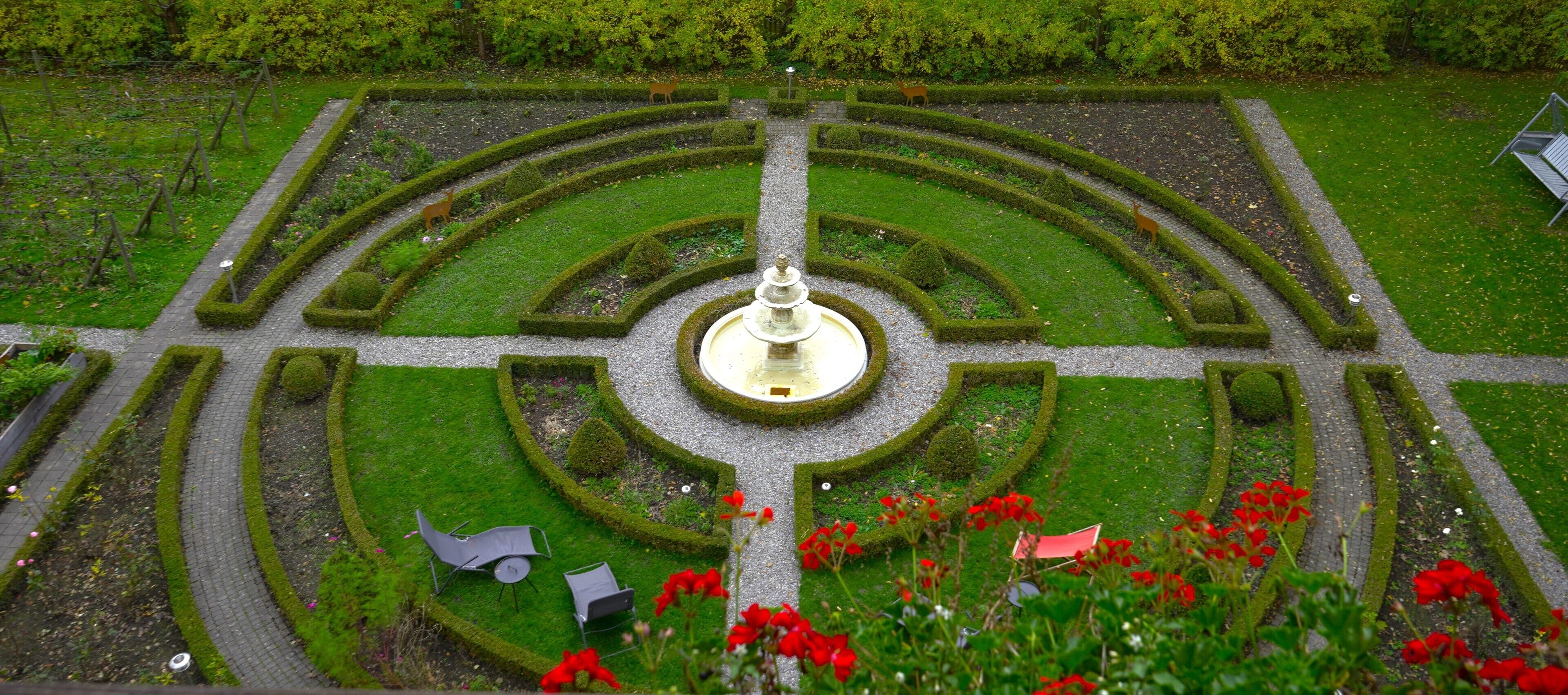 Un jardin à la française avec une fontaine centrale, des haies géométriques et des espaces aménagés pour s’asseoir.