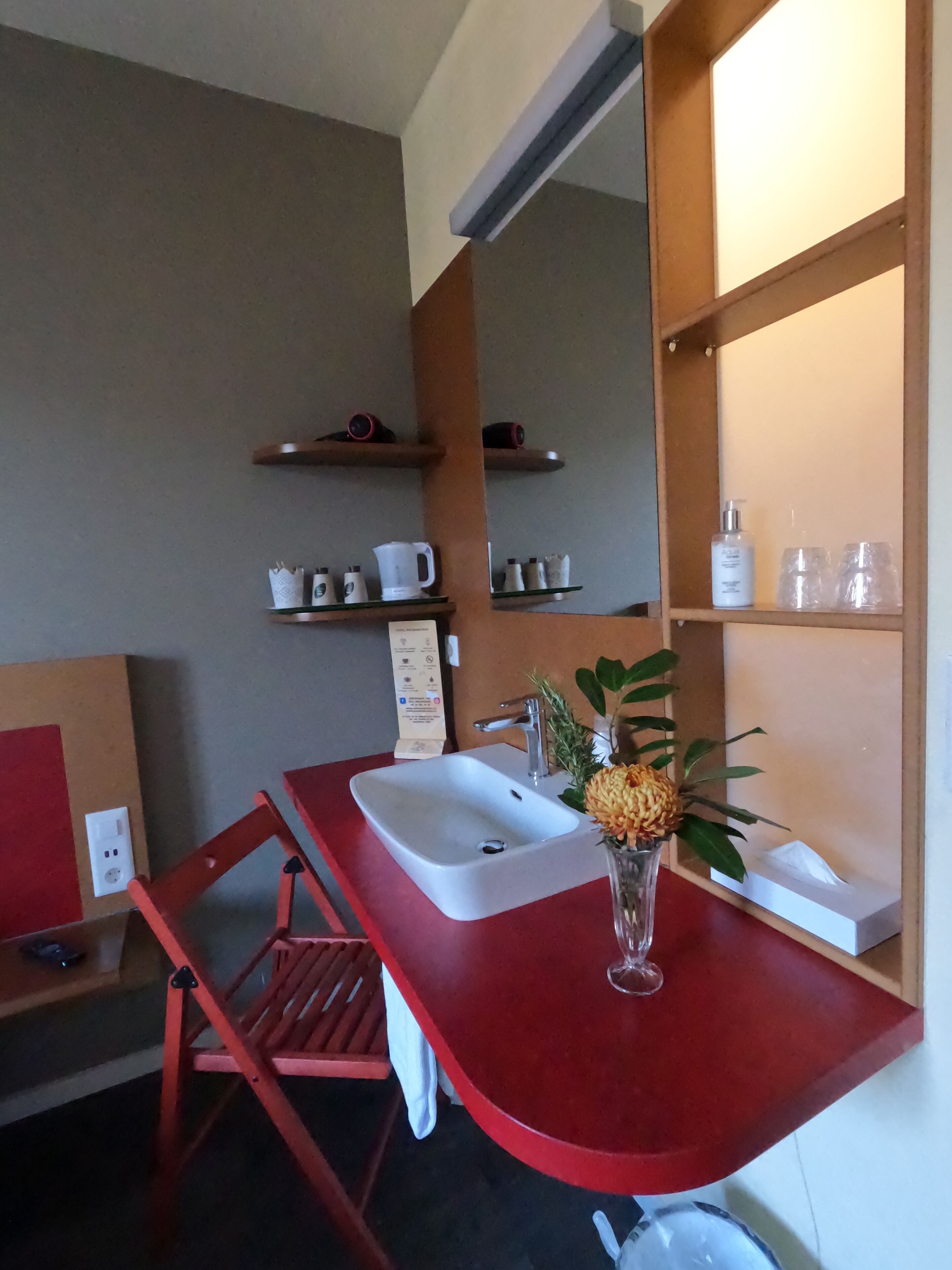 A compact hotel bathroom with a red vanity, a white sink, and a mirror above it. There are shelves with toiletries and a small plant on the vanity.