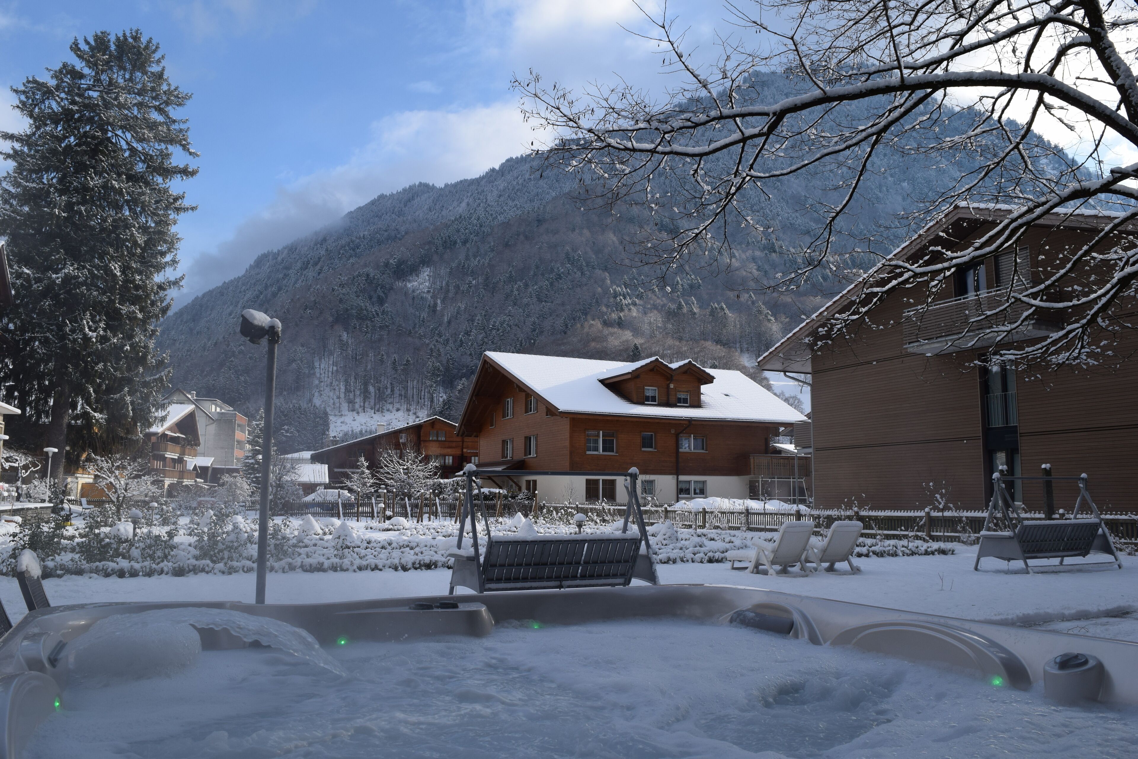 A snow-covered outdoor area with a hot tub, lounge chairs, and a building with a sloped roof.
