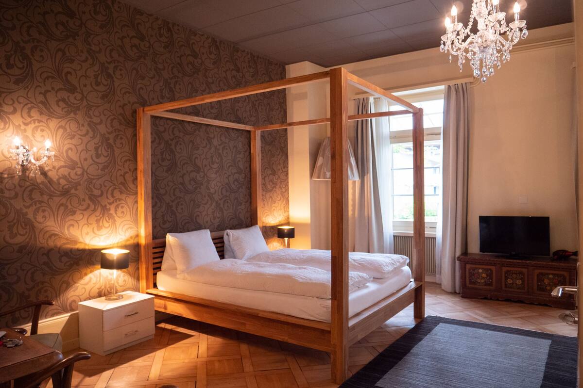 Un lit à baldaquin avec un cadre en bois, une table de chevet avec une lampe, un téléviseur posé sur un meuble et un lustre.