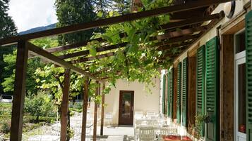 An outdoor patio with white metal chairs and tables, a wooden pergola with vines, and a building with green shutters and a wooden balcony.