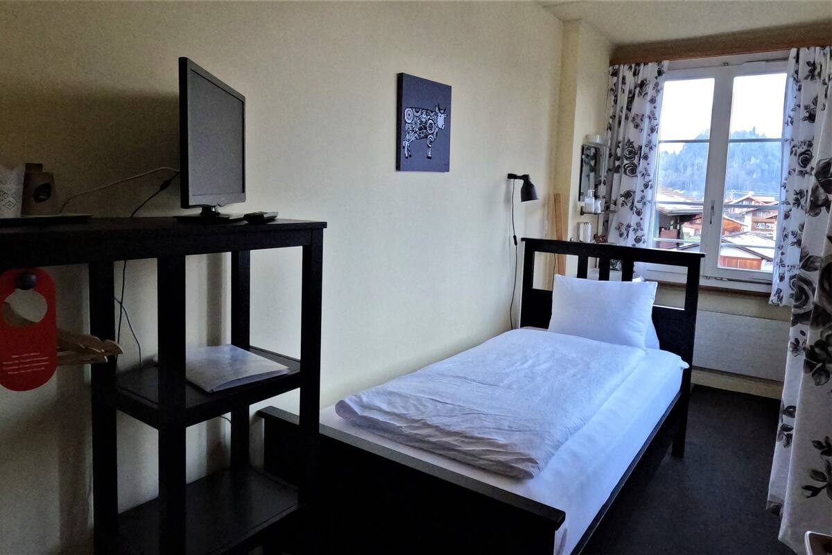 Un lit simple avec un matelas et des oreillers blancs, un bureau avec un écran d’ordinateur, une œuvre d’art représentant un taureau accrochée au mur, et une fenêtre ornée de rideaux à motifs floraux.