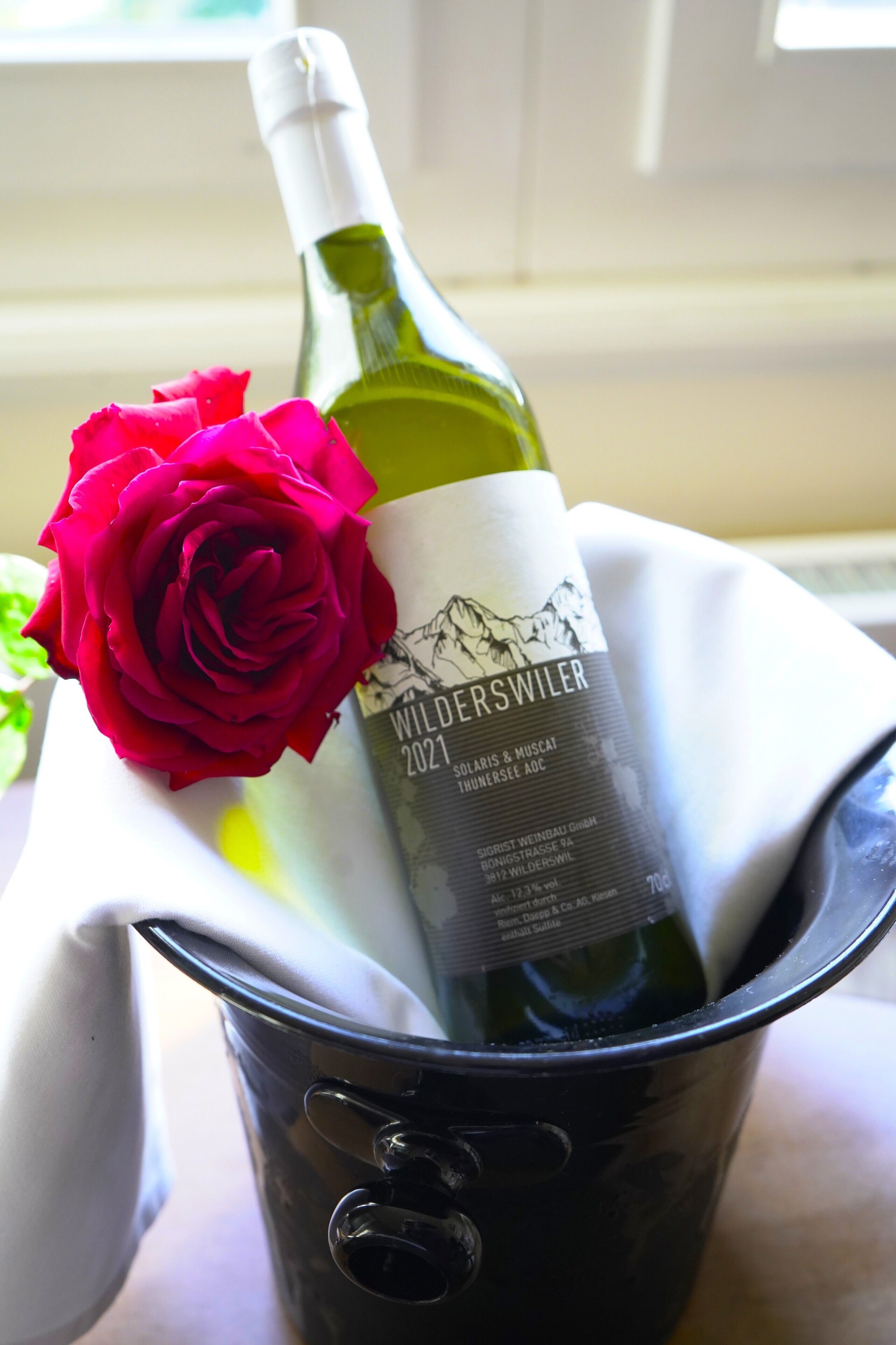 A bottle of wine with a label, a red rose, and a white cloth in a bucket.