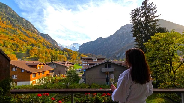 A woman on a balcony overlooking a village with houses and mountains in the background.
