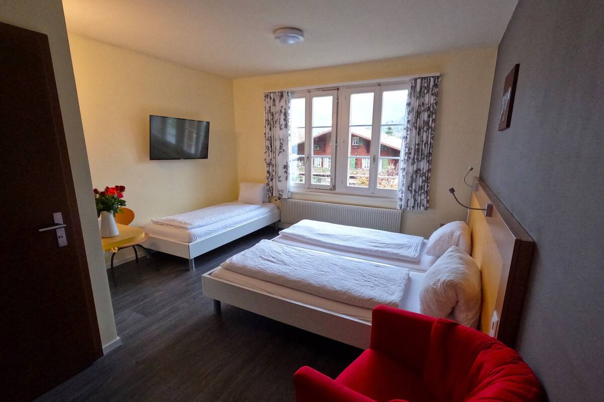 Une chambre d’hôtel avec deux lits simples, un fauteuil rouge, une télévision et une fenêtre avec des rideaux.