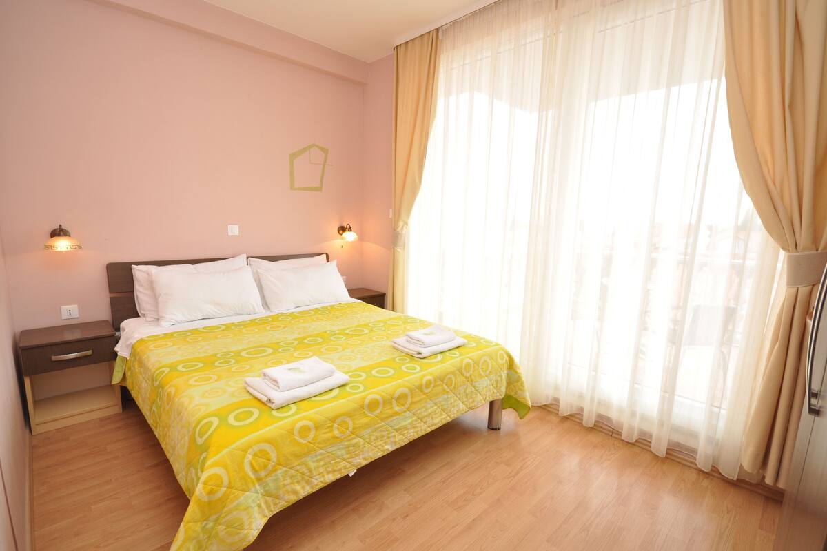 A neatly made bed with a yellow patterned bedspread and white pillows, a bedside table with a lamp, and a window with sheer curtains.
