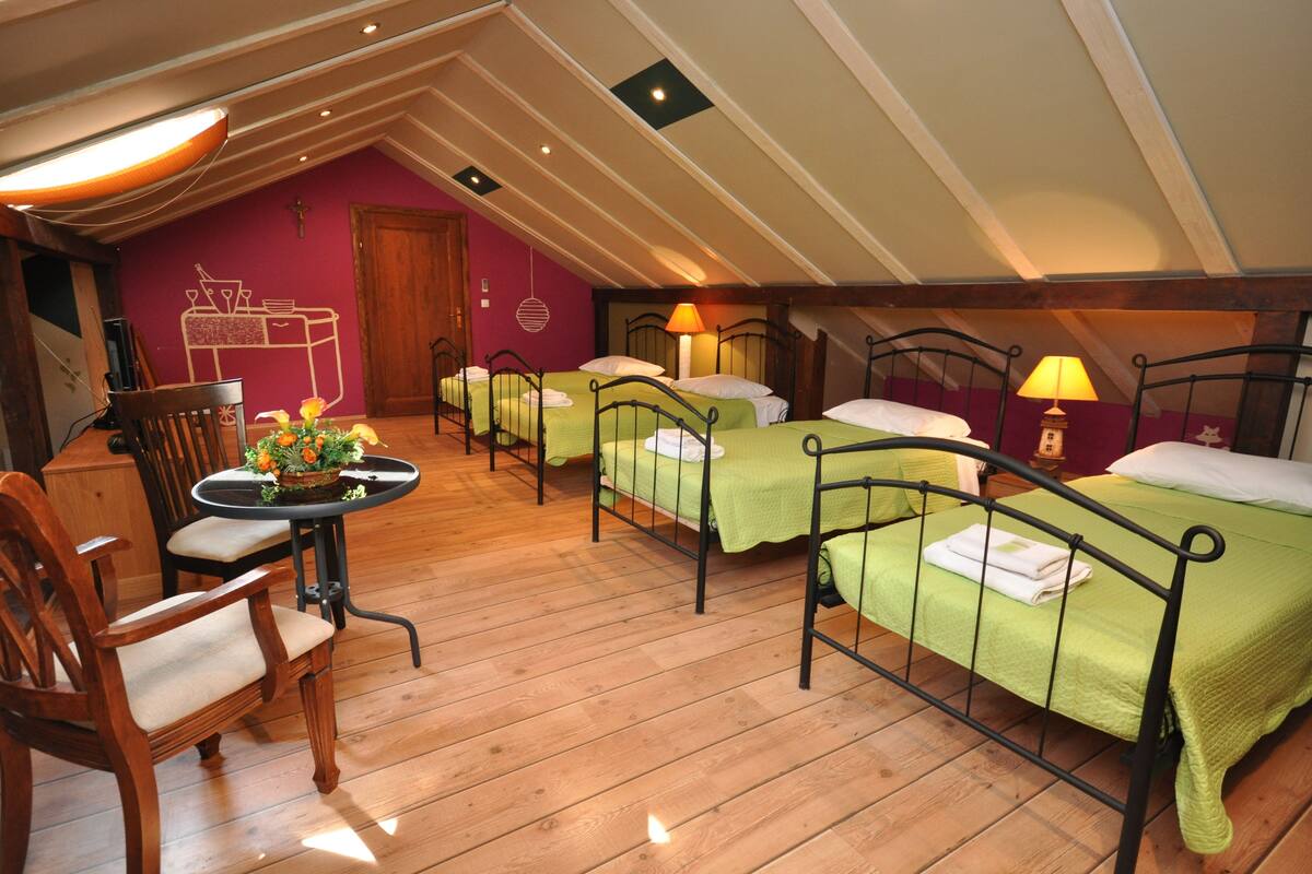 A dormitory room with four beds, wooden floors, a dining table with chairs, and a small table with a flower arrangement.