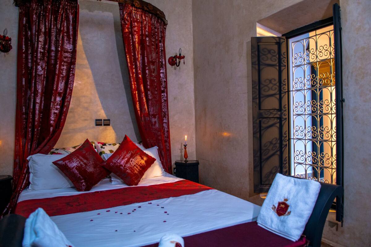 A bed with red and white bedding, a nightstand with a candle, and a window with decorative iron bars.