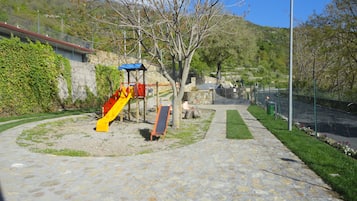 Explore the playground with a slide and climbing structures on a paved surface, surrounded by a hedge and grassy area.