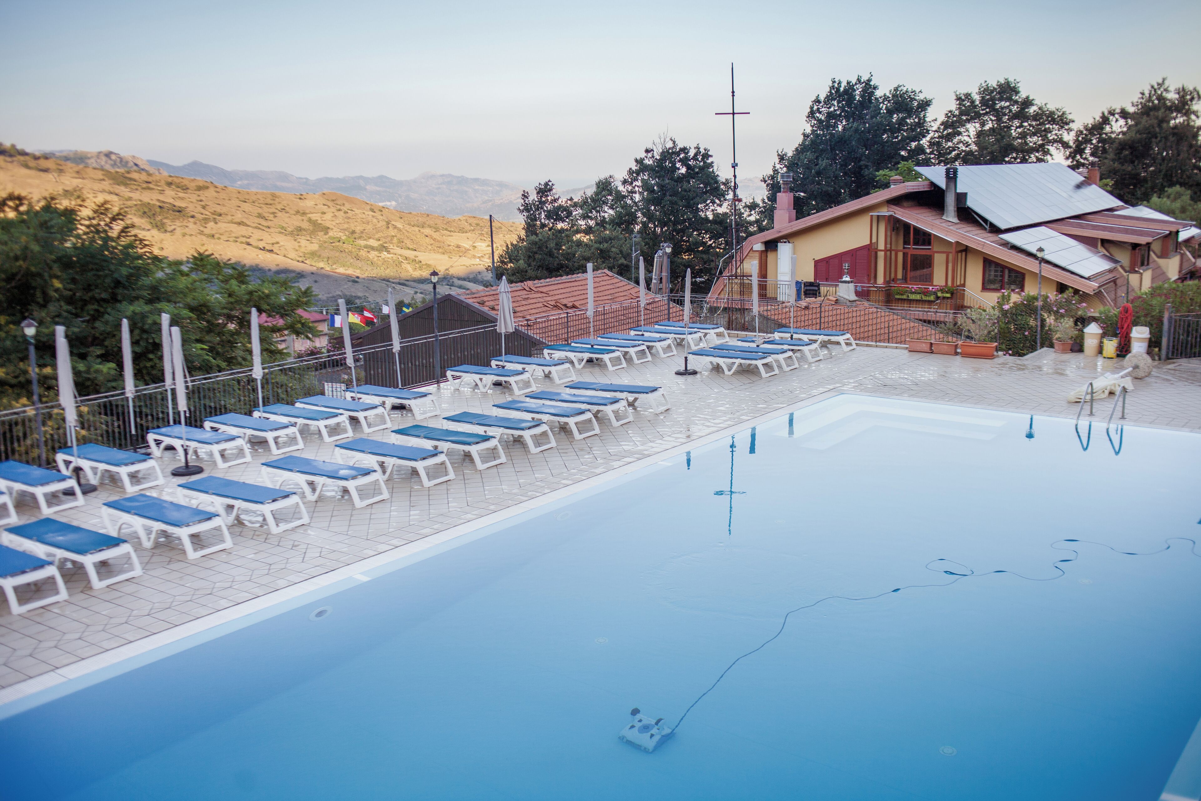 A swimming pool with lounge chairs, a building with a sloped roof, and a mountainous backdrop.