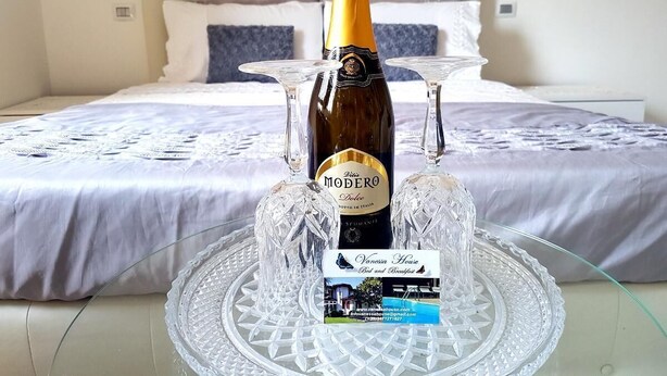 Uma cama com cabeceira branca, uma garrafa de vinho e duas taças de vinho sobre um jogo de copos de vidro.