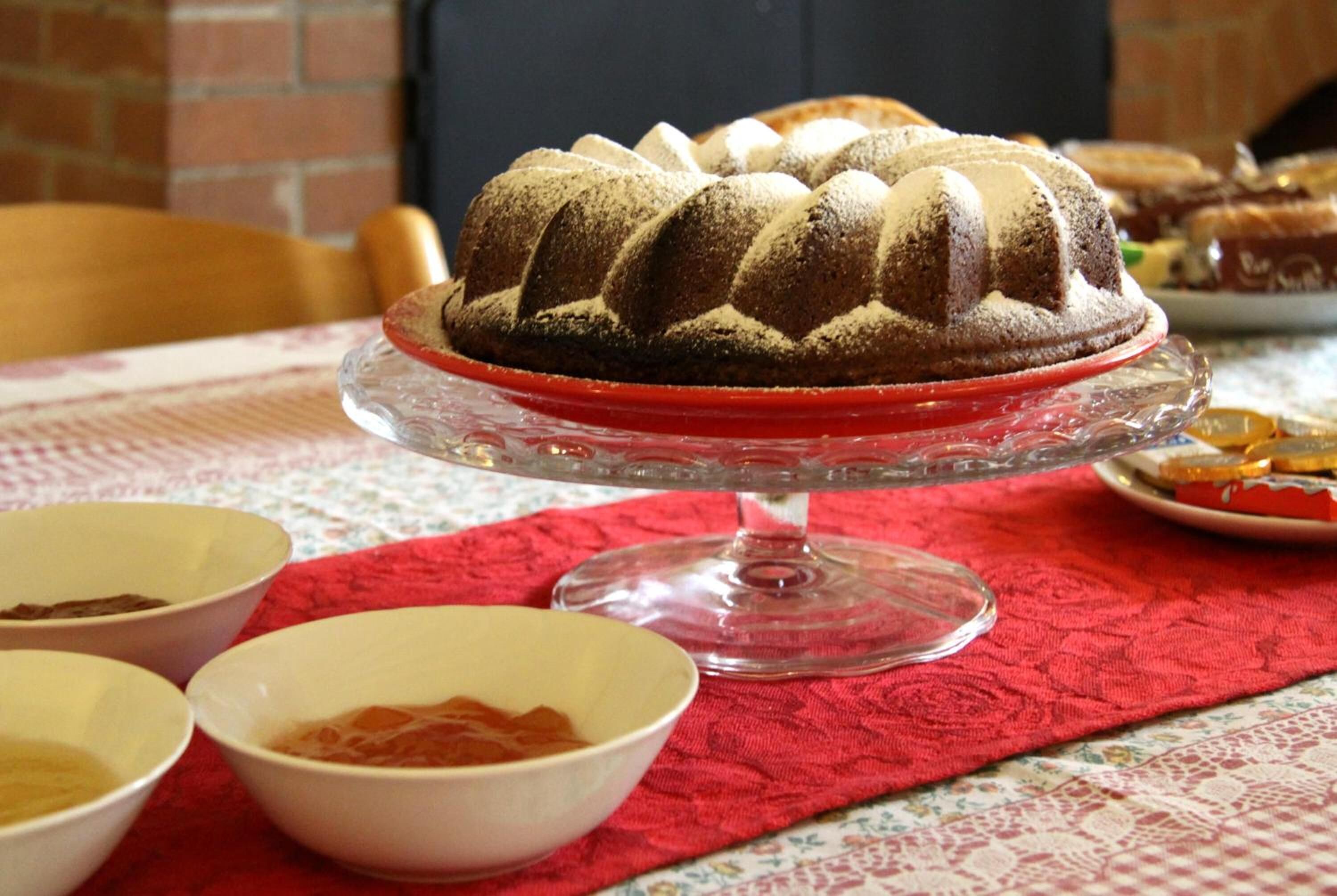Um bolo bundt em um suporte de vidro com um tecido vermelho por baixo, rodeado por tigelas sobre uma toalha de mesa.