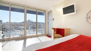 Habitación de hotel con un ventanal grande, una cama con una manta roja, un pequeño escritorio de madera y un televisor montado.