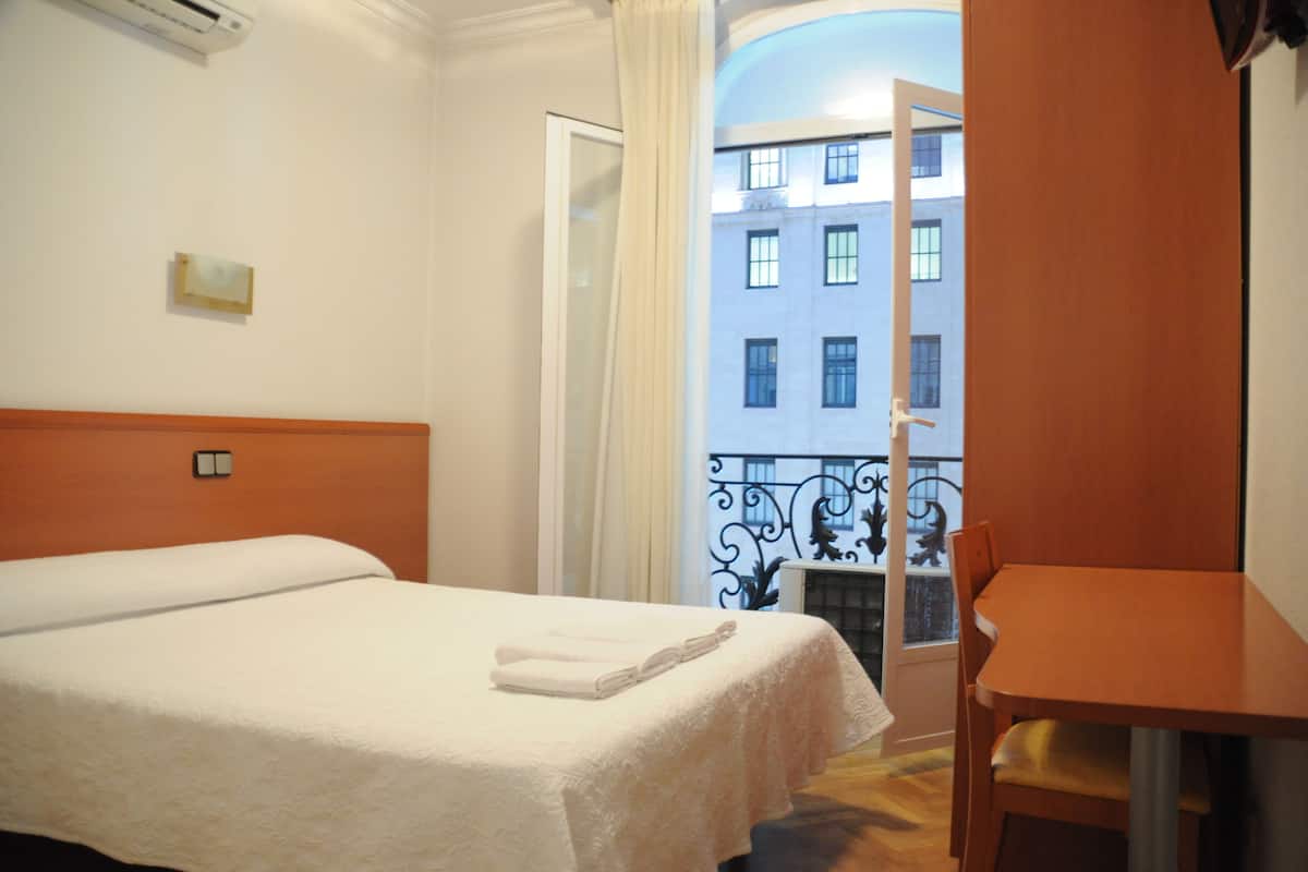 A hotel room with a bed, a wooden table, and a view of a building through an open door.