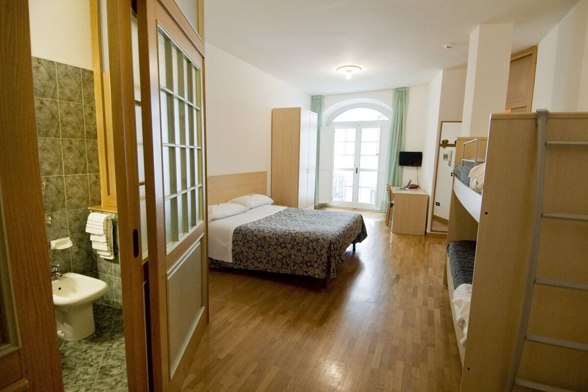 Habitación de hotel con suelo de madera, cama con edredón estampado, mesita de noche con lámpara, televisor y armario.