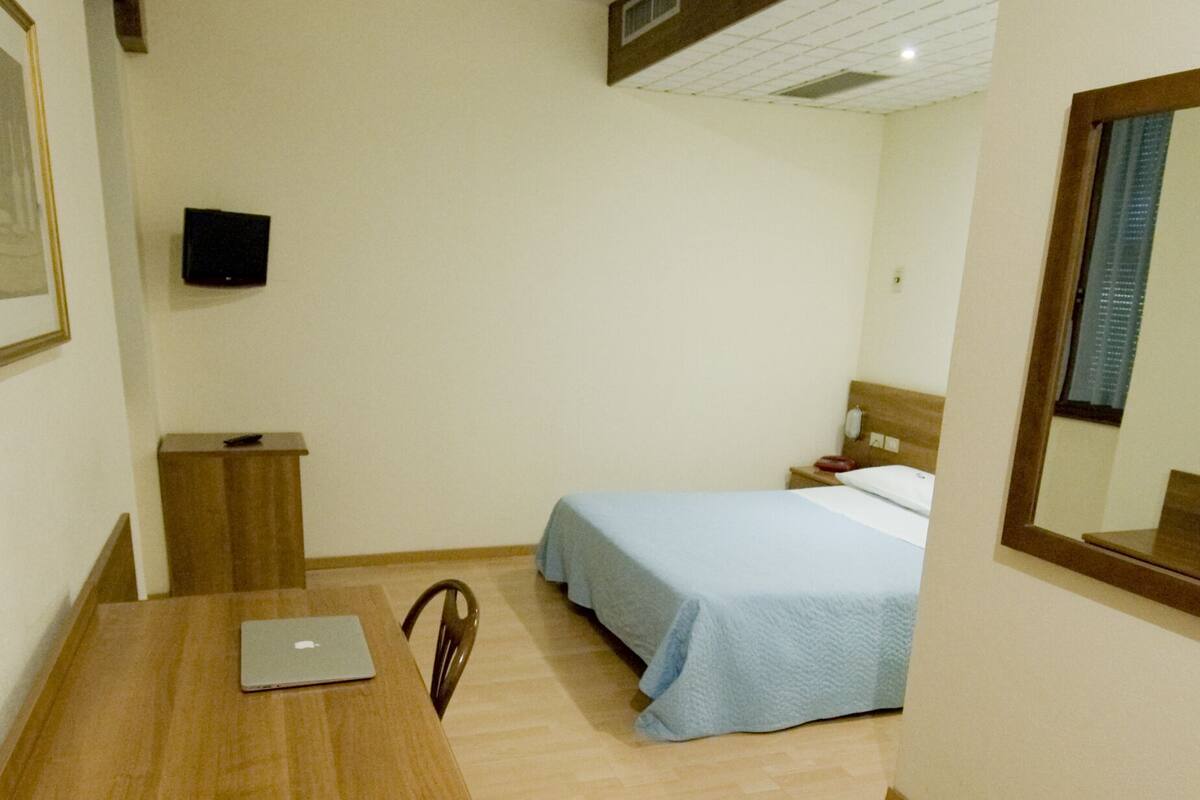 Un dormitorio con una cama, un escritorio con una computadora portátil, una silla, una mesita de noche y un televisor en la pared.
