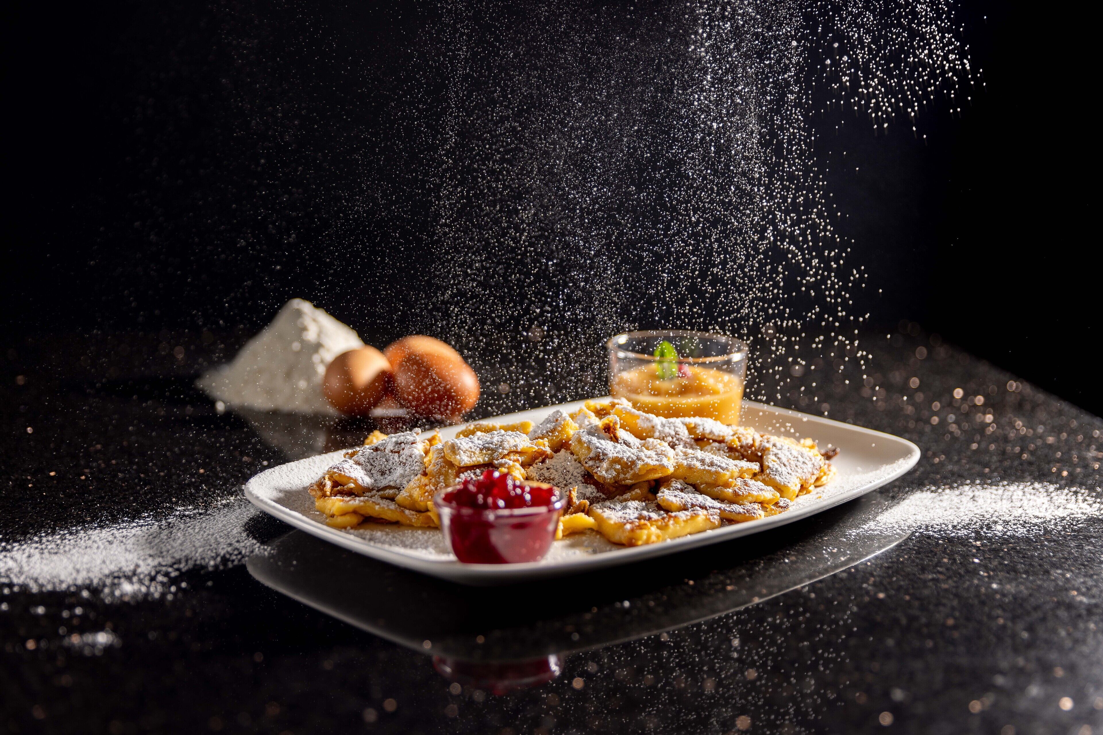 A dessert plate with powdered sugar, a bowl of red sauce, and a glass of drink on a dark surface.
