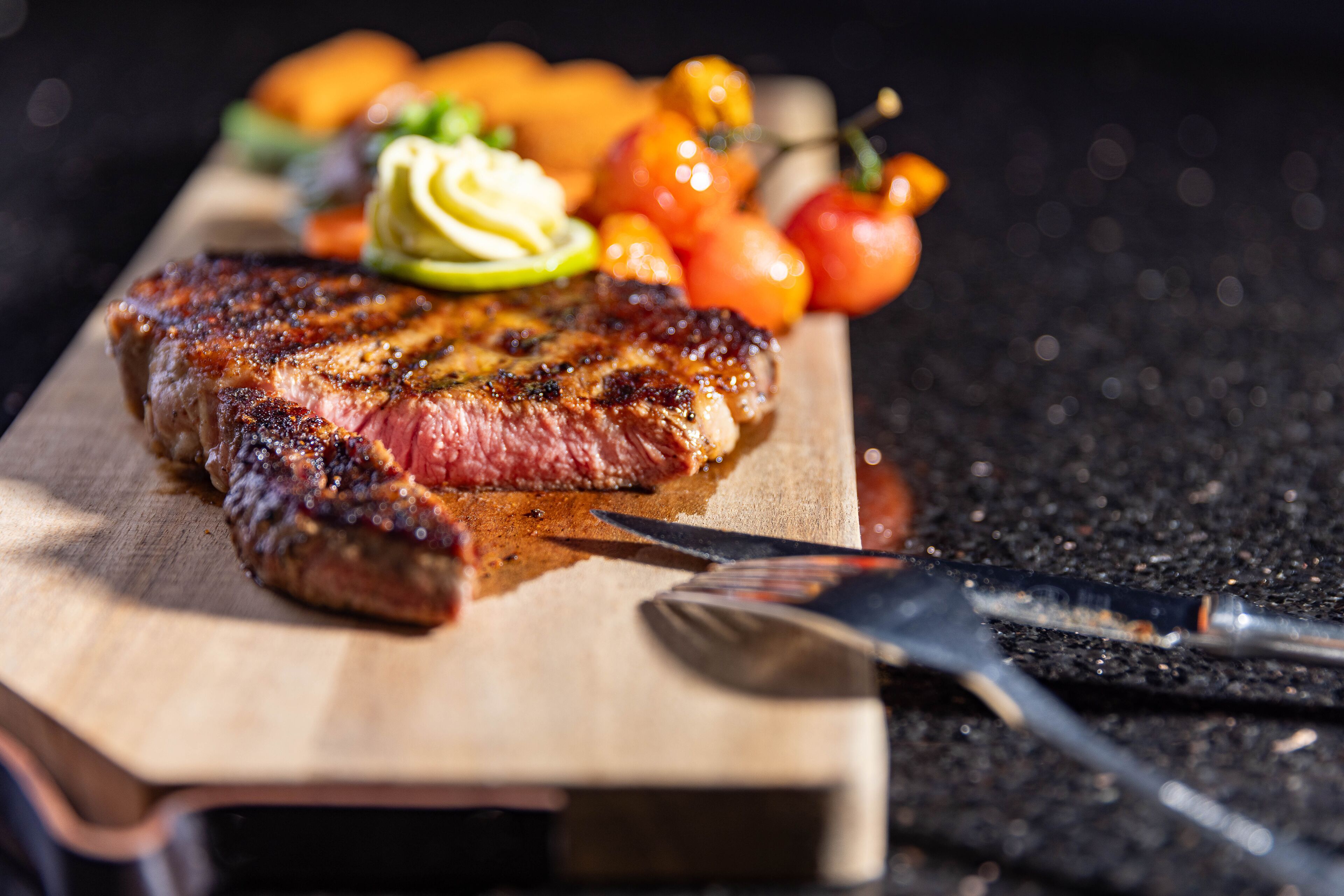 Een steak met mes en vork op een houten snijplank, vergezeld van gegrilde groenten en een kleine salade.