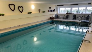 An indoor swimming pool with a tiled floor, surrounded by chairs and tables, and decorated with heart-shaped wall art. - Hotel am Wald (Bad Toelz)