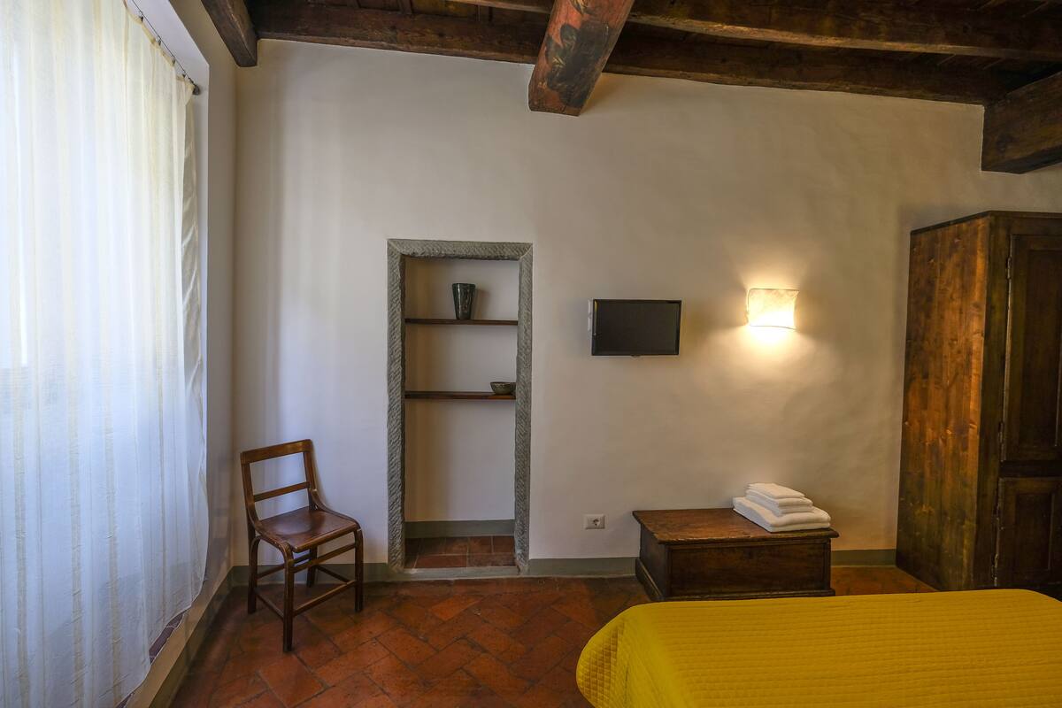 A room with a bed, a chair, a shelf with a cup, a wall-mounted light, and a wooden door.