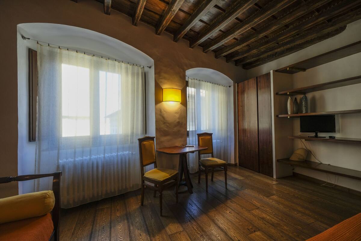 A cozy room with wooden flooring, a round table with chairs, a TV on a shelf, and a lamp on the wall.