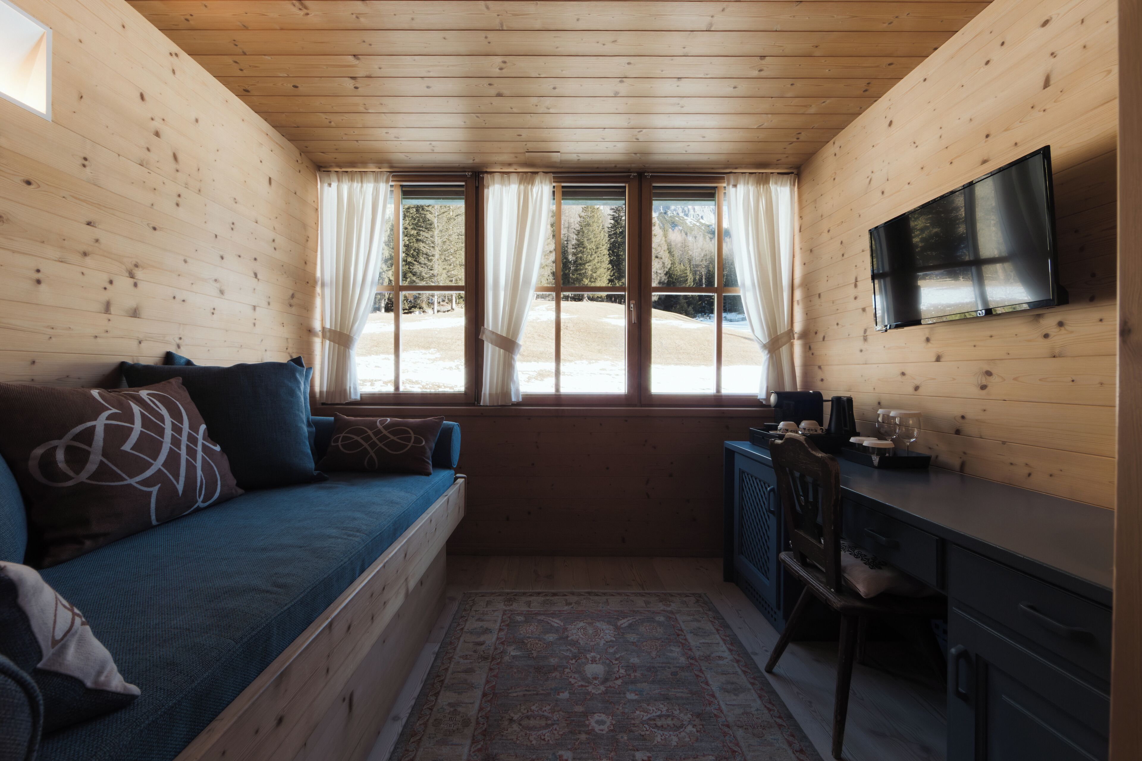 A cabin interior with a bed, a desk, a television, and a window with curtains.