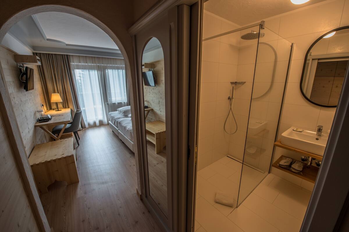 A hotel room with a wooden floor, a bed, a desk with a chair, a TV, a shower area with a glass door, and a bathroom with a sink and mirror.