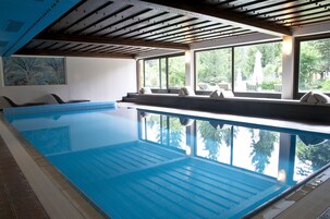 Un'ampia piscina interna con acqua cristallina, travi di legno al soffitto e grandi finestre che offrono vista sul verde circostante.