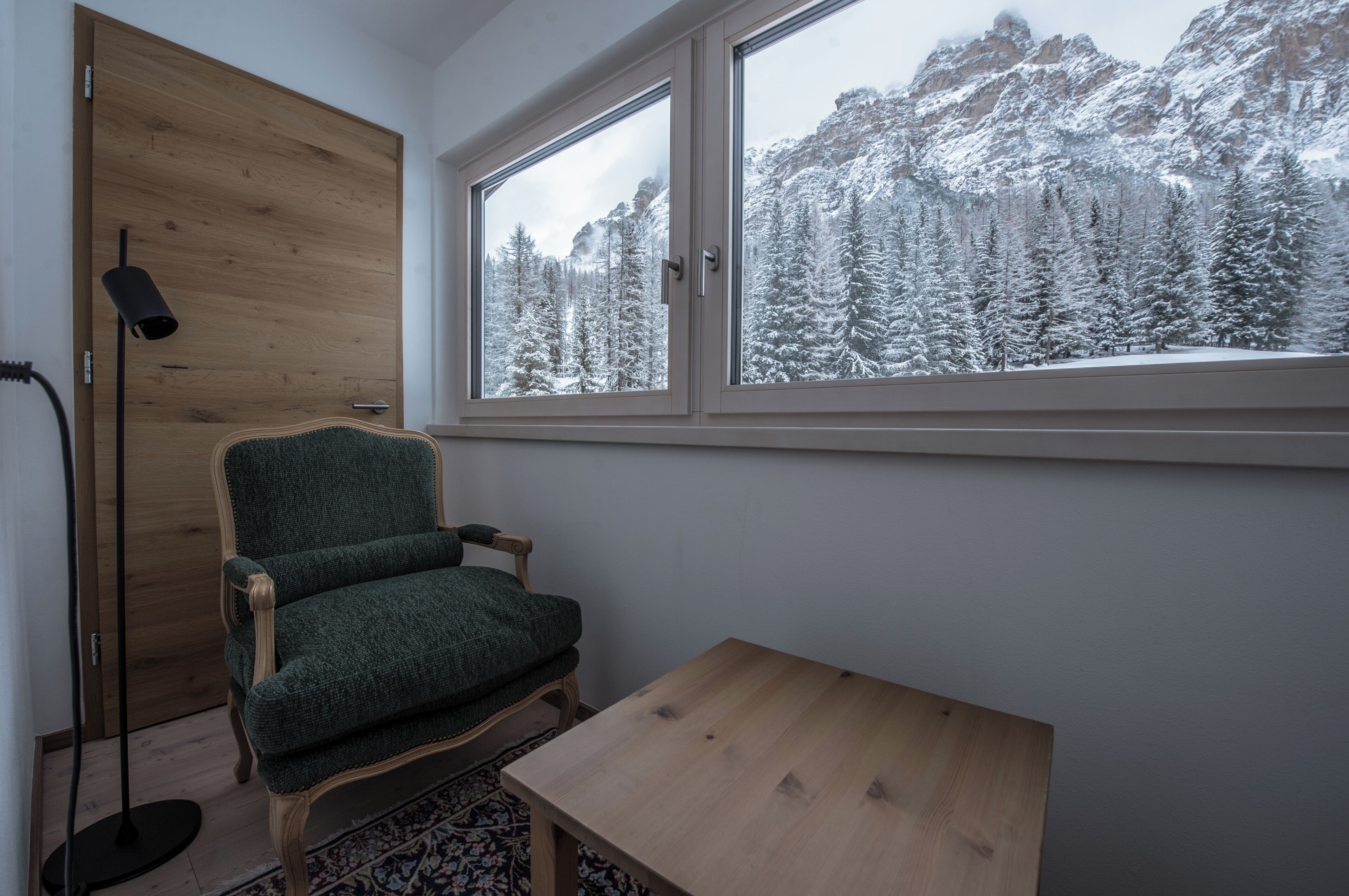 Ein Zimmer mit Holztür, einem grünen Polsterstuhl, einem kleinen Holztisch und einem Fenster mit Blick auf einen verschneiten Berg.