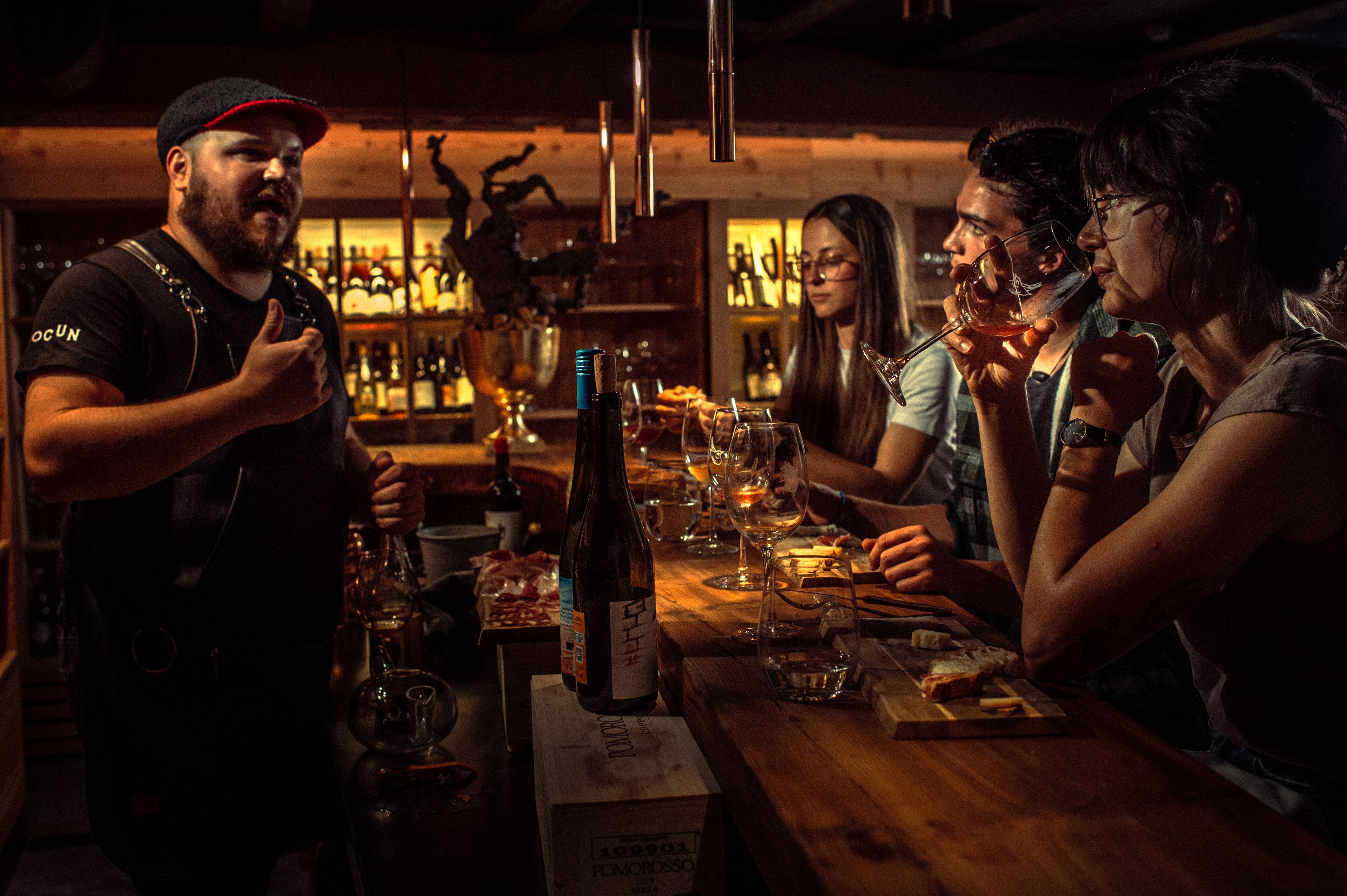 A bar setting with a wooden table, bottles, glasses, and a person standing.