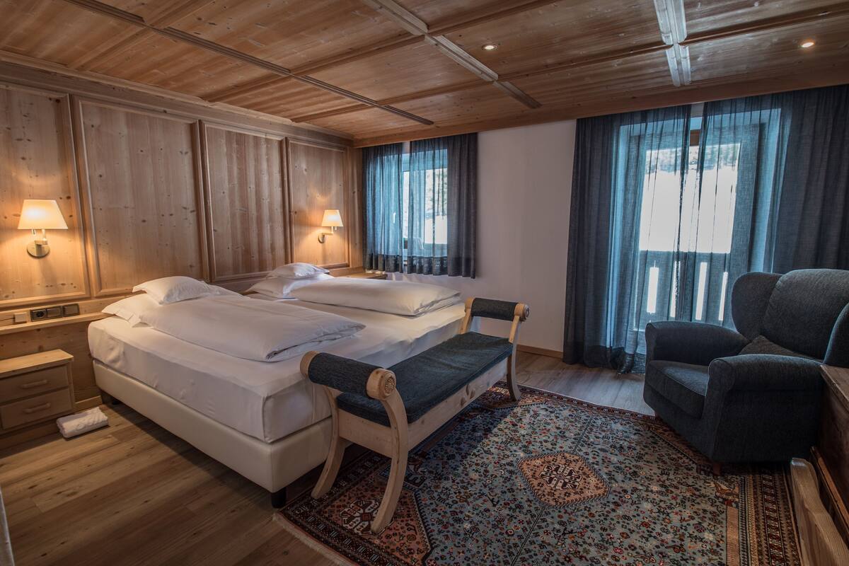 A hotel room with two beds, a bench, a chair, and a patterned rug.