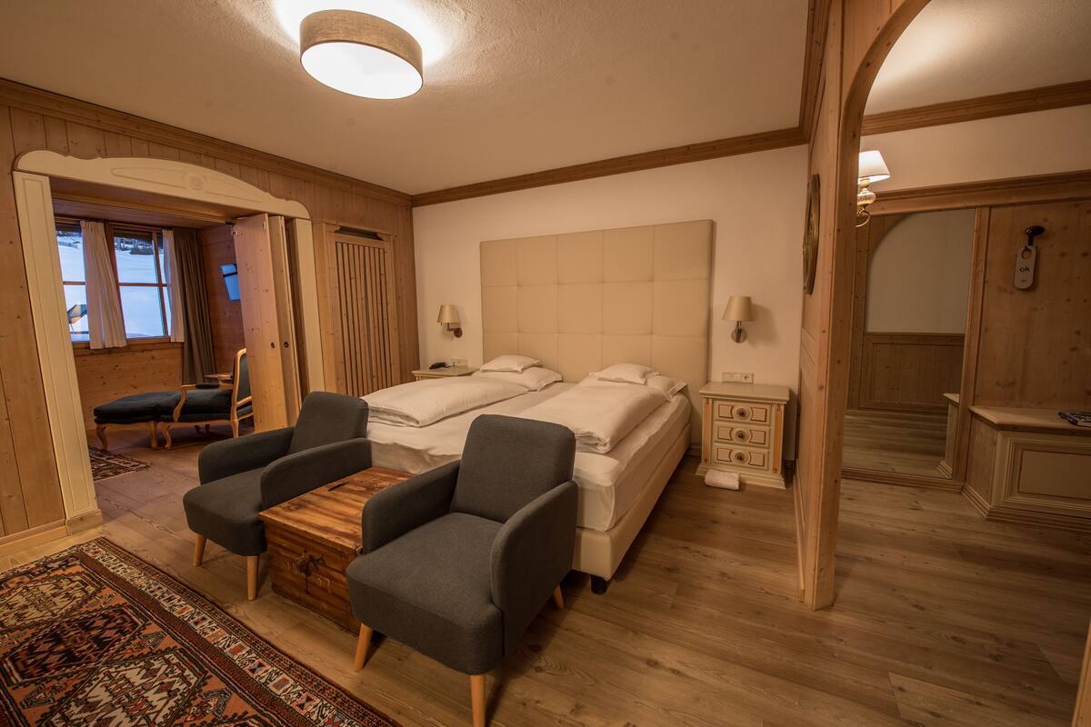 A bedroom with two beds, a wooden floor, a desk with chairs, and a window with curtains.