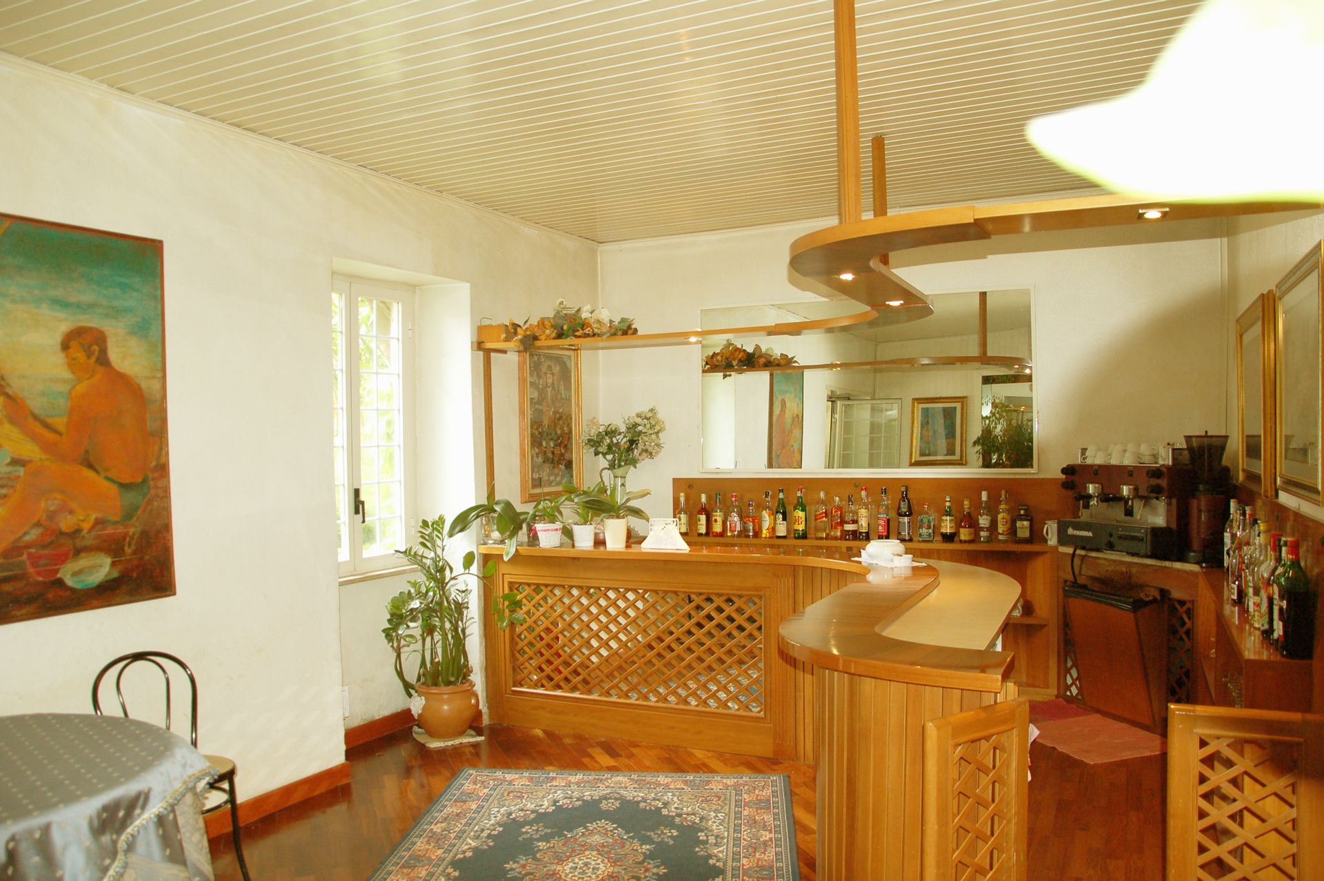 Una habitación con barra de madera, zona de comedor con mesa y sillas, un cuadro grande en la pared y una planta en maceta.