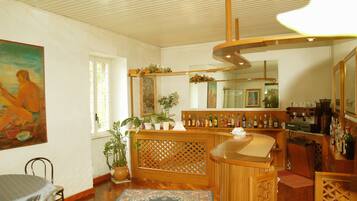 Una habitación con barra de madera, zona de comedor con mesa y sillas, un cuadro grande en la pared y una planta en maceta.