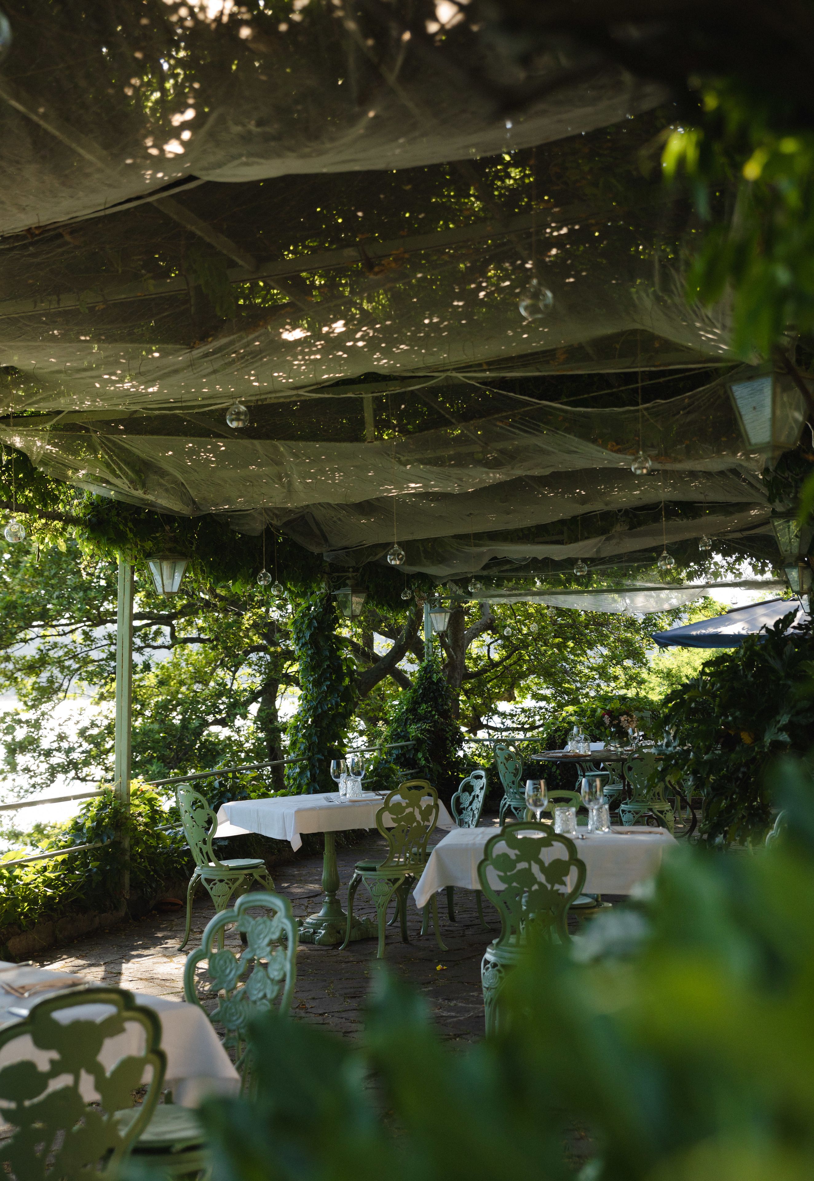 An outdoor dining area with tables and chairs under a canopy, surrounded by greenery.