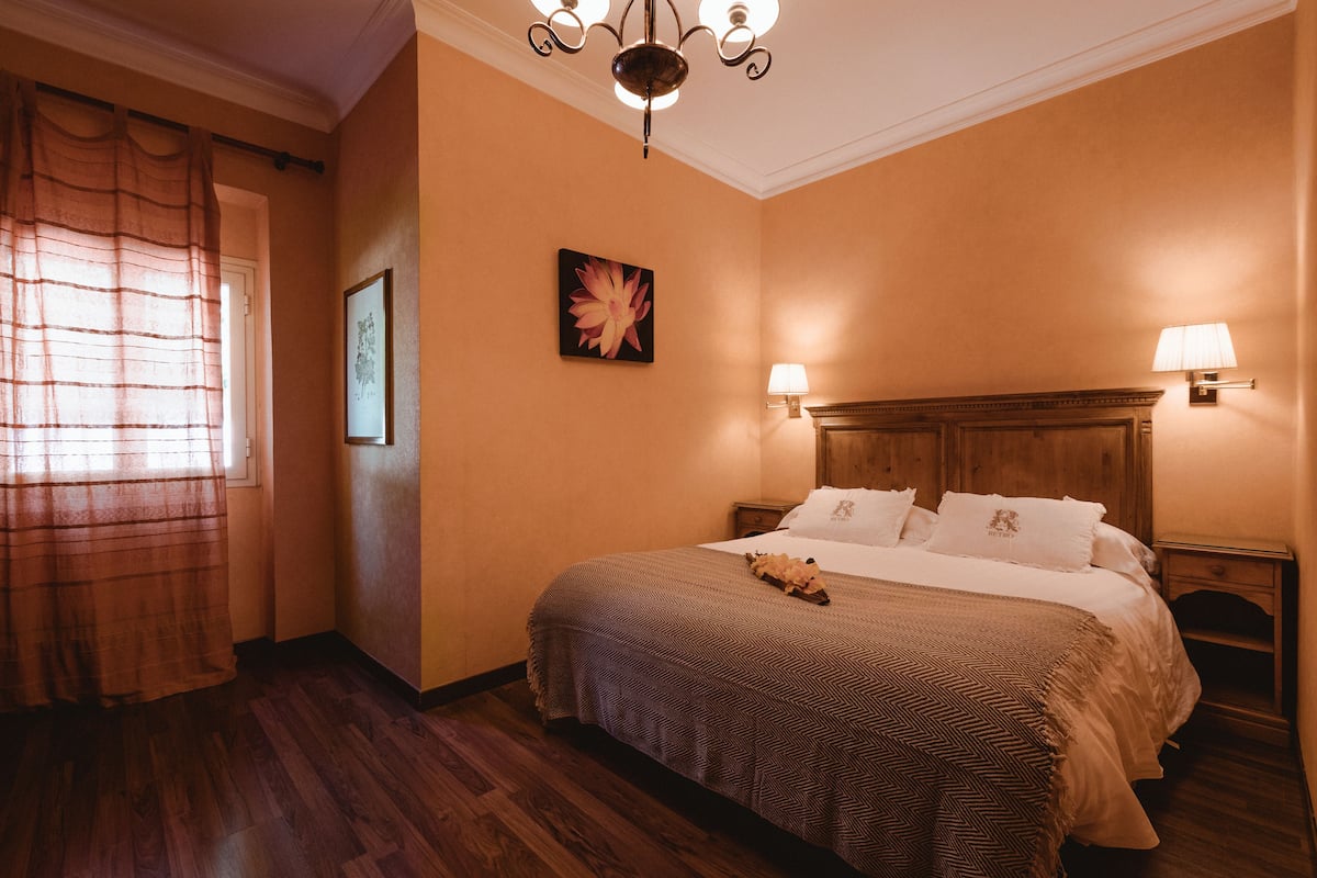 Habitación de hotel con cabecera de madera, una cama grande con colcha estampada, dos lámparas de noche, un cuadro en la pared y una ventana con cortinas.