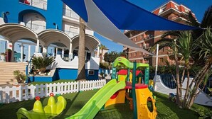 A children's play area with slides and climbing structures, surrounded by a white picket fence and palm trees. - Ambassador (Tortoreto)